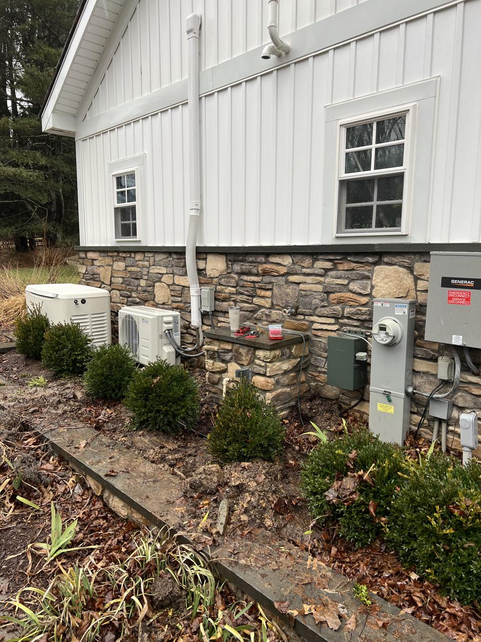 A Generac standby generator, mini split and Generac transfer switch beside a white house with a stone base